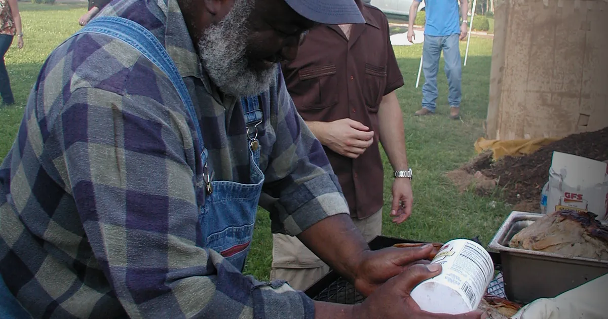 A pair of hands with a mop basting a whole hog over a long pit of glowing coals, smoke rising into the dark