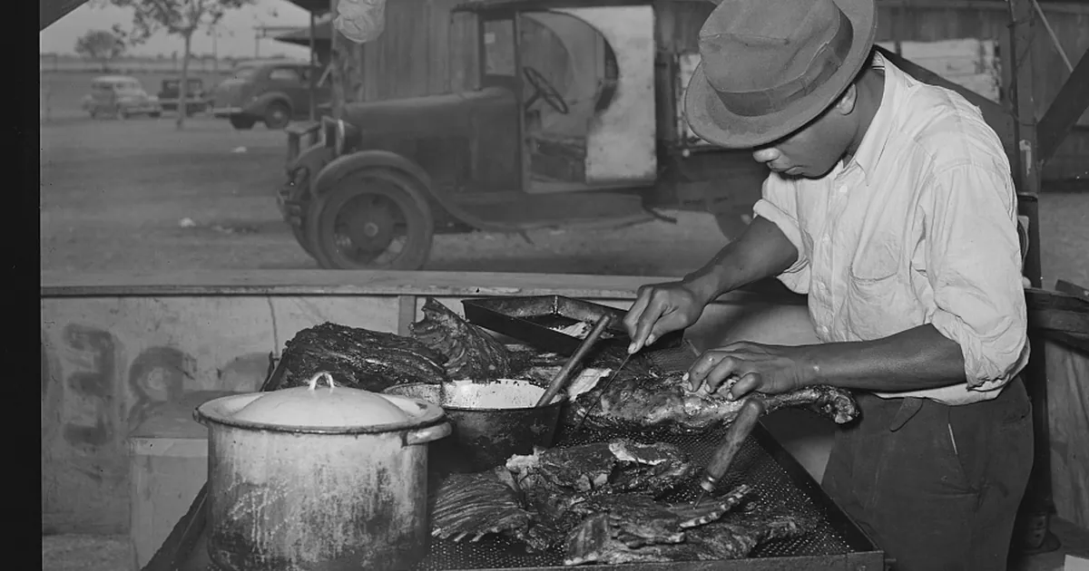 Black-and-white photograph from the 1930s of cooks in white aprons and caps working around a brick-lined barbecue pit, tubs of barbecued beef beside them and a crowd gathered behind